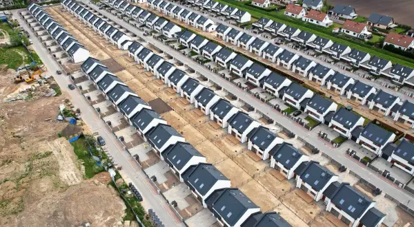 Nowe osiedle domów jednorodzinnych, przykład suburbanizacji. Domy szeregowe z ciemnymi dachami i białymi elewacjami, otoczone placem budowy.