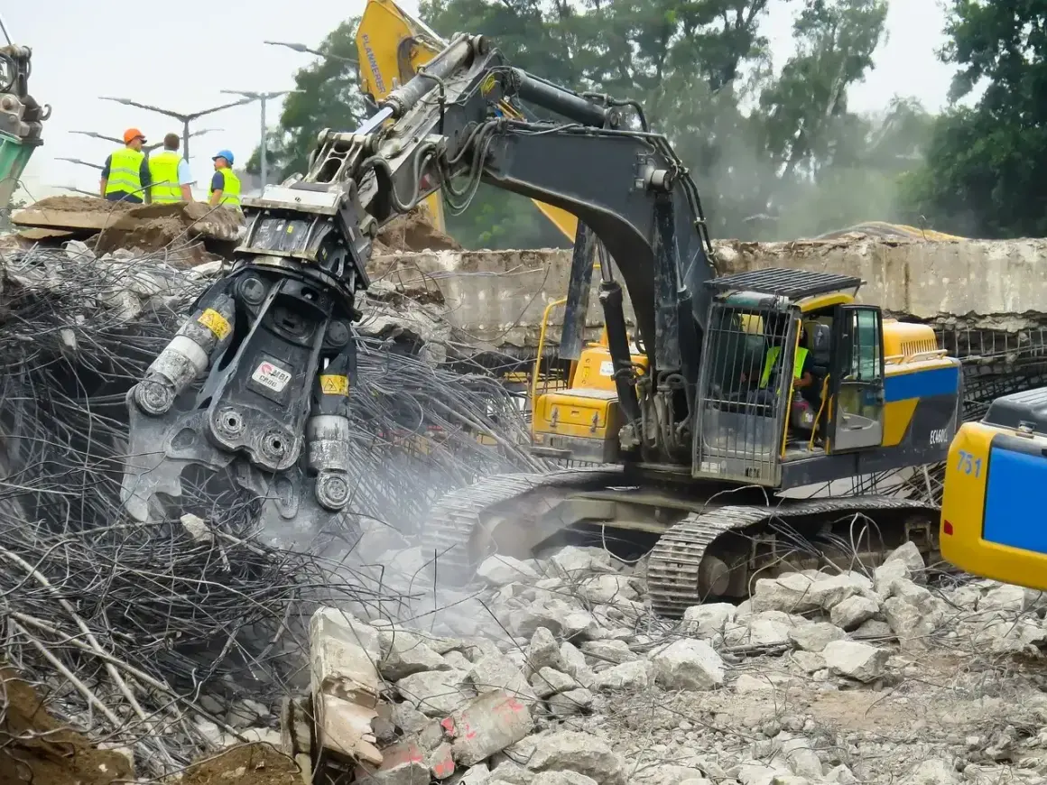 Maszyna budowlana wykonuje rozbiórkę, krusząc beton i stal. W tle widać pracowników w kamizelkach.