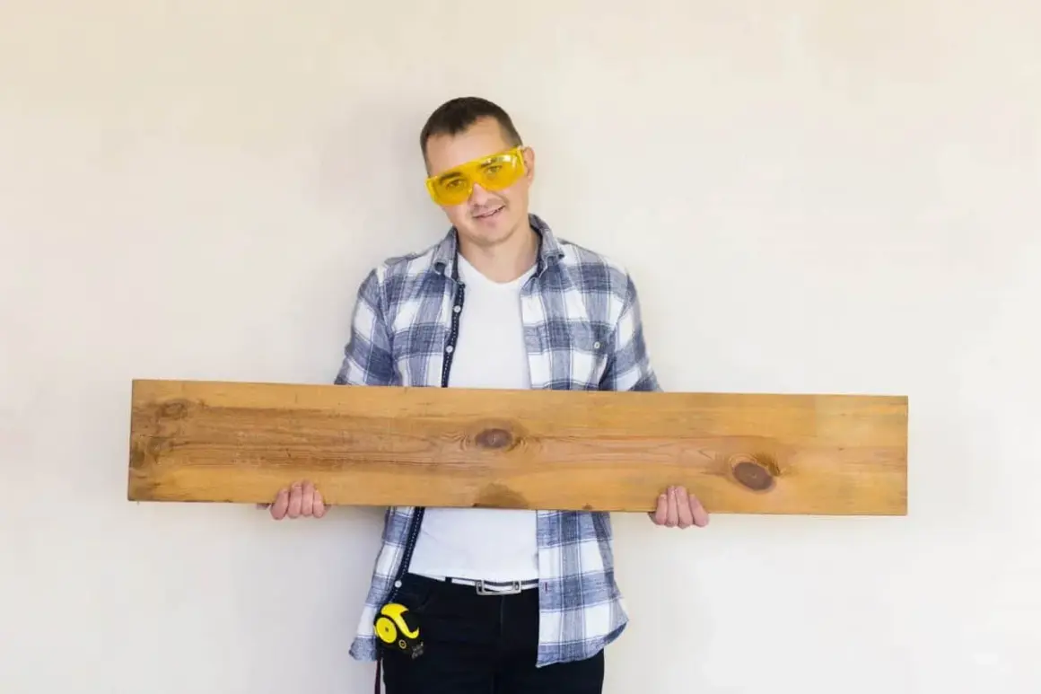 Mężczyzna w okularach ochronnych trzyma drewnianą deskę, która może posłużyć jako panele podłogowe na ścianie.