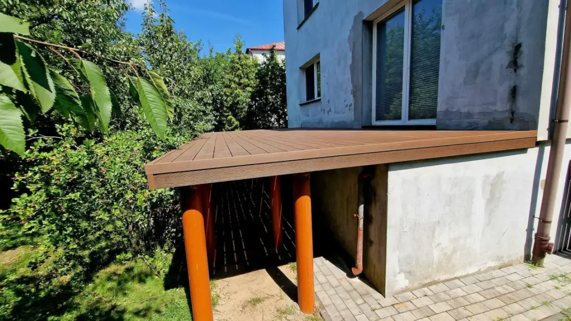 Nowy taras na słupach betonowych, z brązową, drewnopodobną nawierzchnią, otoczony zielenią.