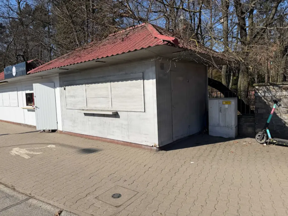 Mały lokal użytkowy z czerwoną, falistą dachem i białymi okiennicami. Obok stoi hulajnoga elektryczna.