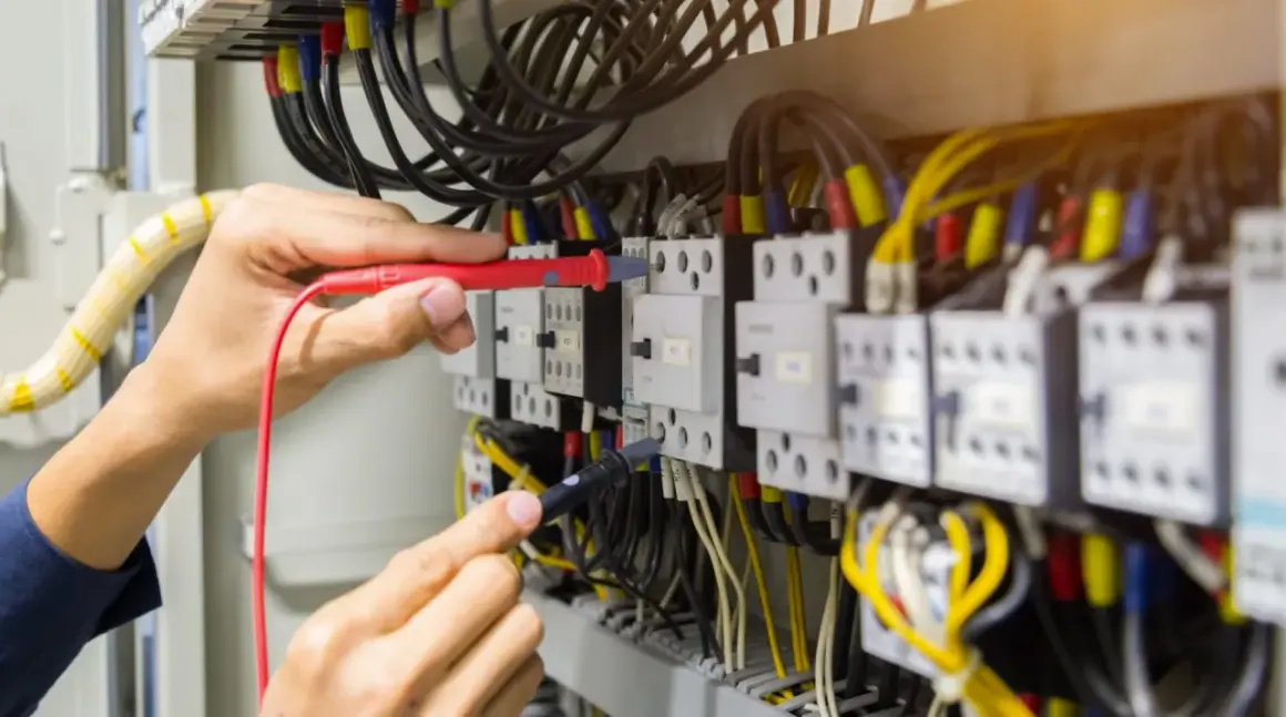 Elektryk sprawdza przewody w szafie sterowniczej. Widać multimetr i liczne kable, w tym przewód neutralny.