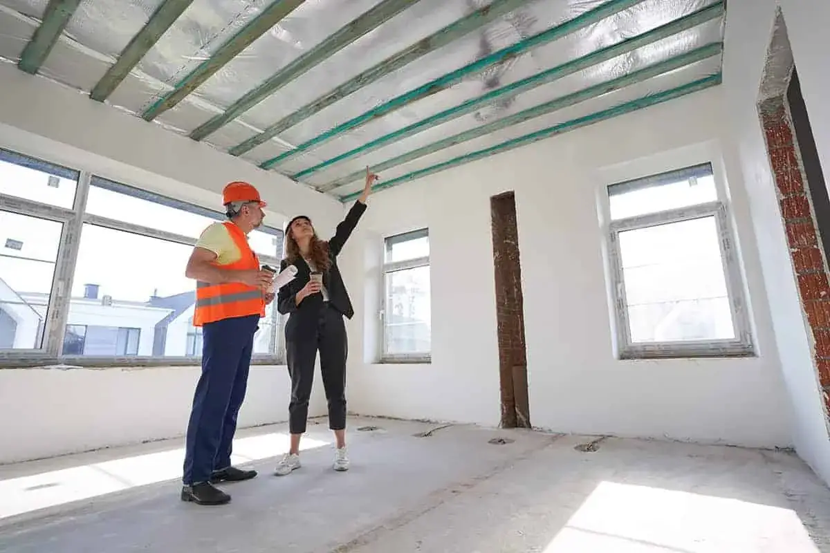Inżynier i klientka omawiają sufity podwieszane, cena za 1m2 robocizna. Wnętrze w stanie surowym, widoczne belki stropowe.