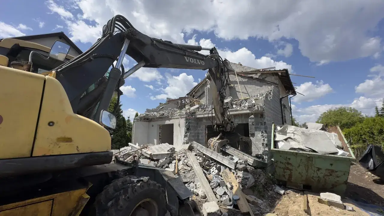 Koparka Volvo pracuje przy rozbiórce domu. Gruz piętrzy się wokół, a kontener czeka na załadunek.