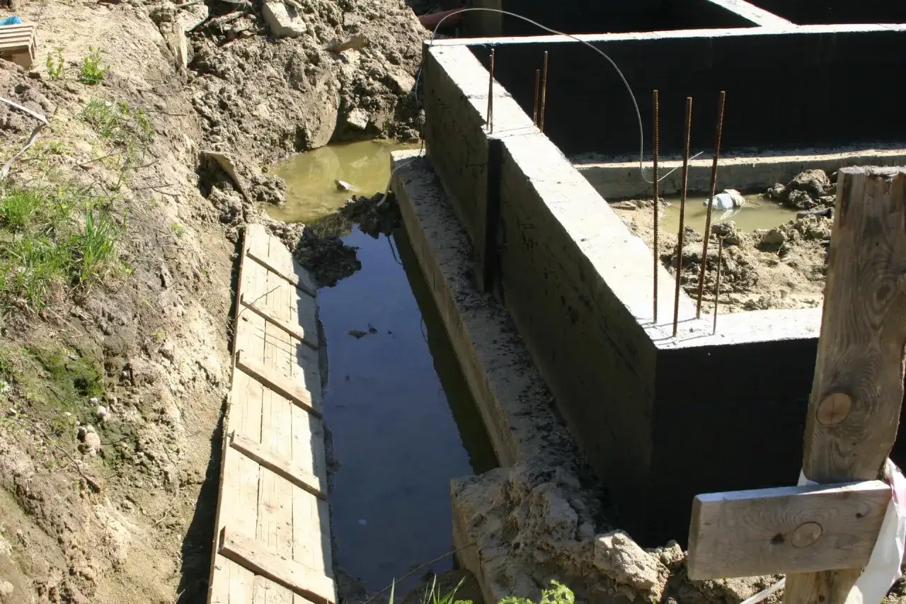 Wykop pod fundamenty wypełniony wodą, co wskazuje na wysoki poziom wód gruntowych. Betonowe ściany fundamentowe są już częściowo wykonane.