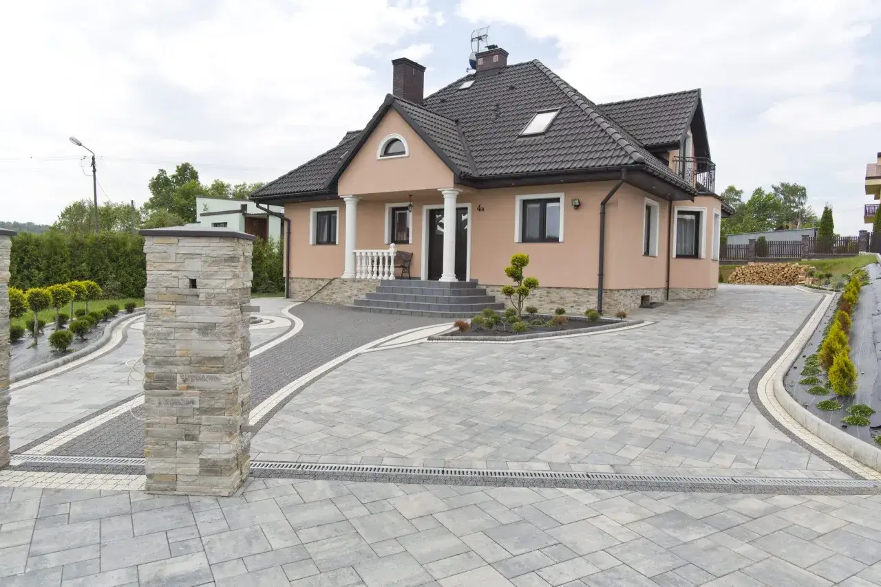 Elegancki podjazd do domu z kostki brukowej, prowadzący do jasnoróżowego budynku z białymi kolumnami.