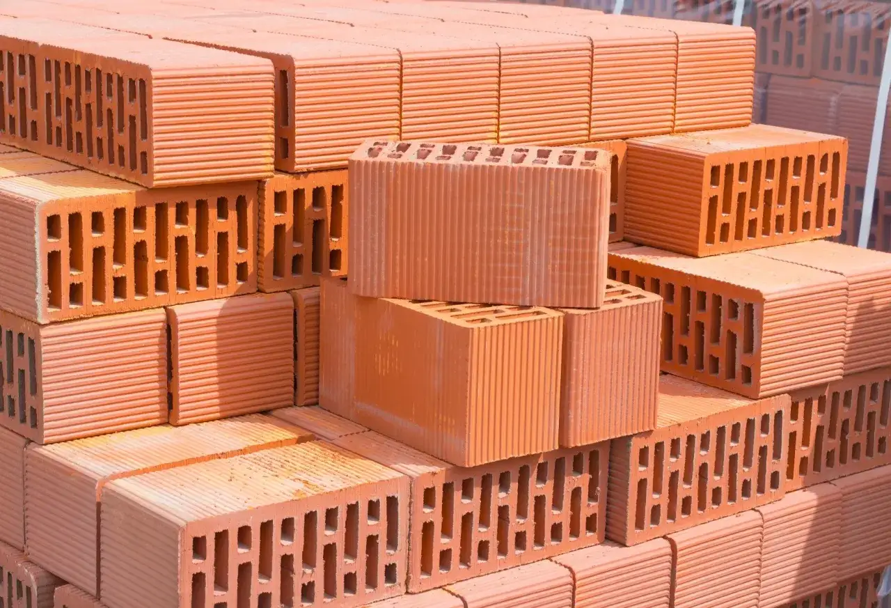 Stos różnych rodzajów cegieł ceramicznych, gotowych do budowy. Widać cegły perforowane i pełne, z charakterystycznymi rowkami.