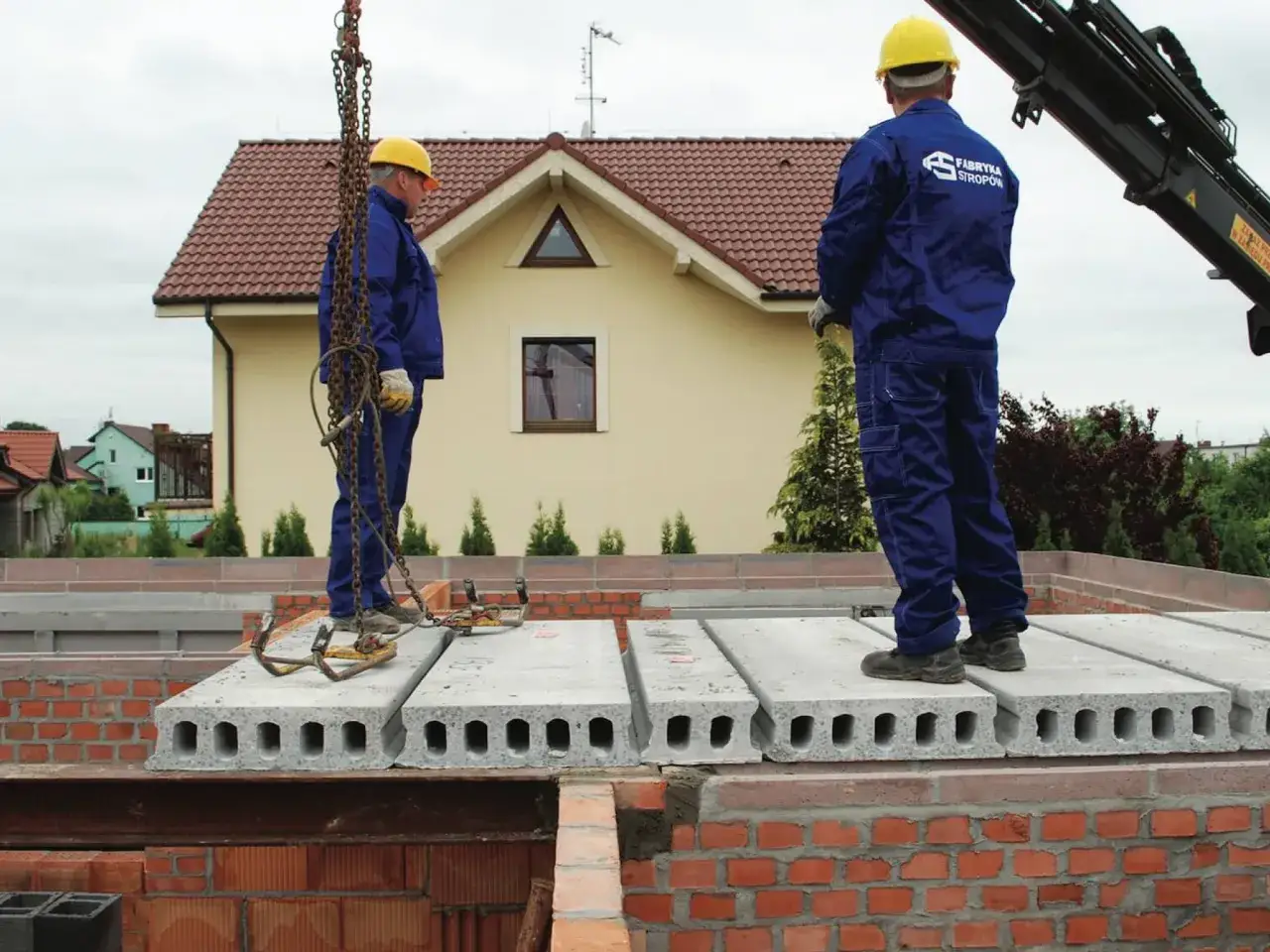 Dwóch robotników w żółtych kaskach i niebieskich kombinezonach montuje strop filigran na budowie domu.