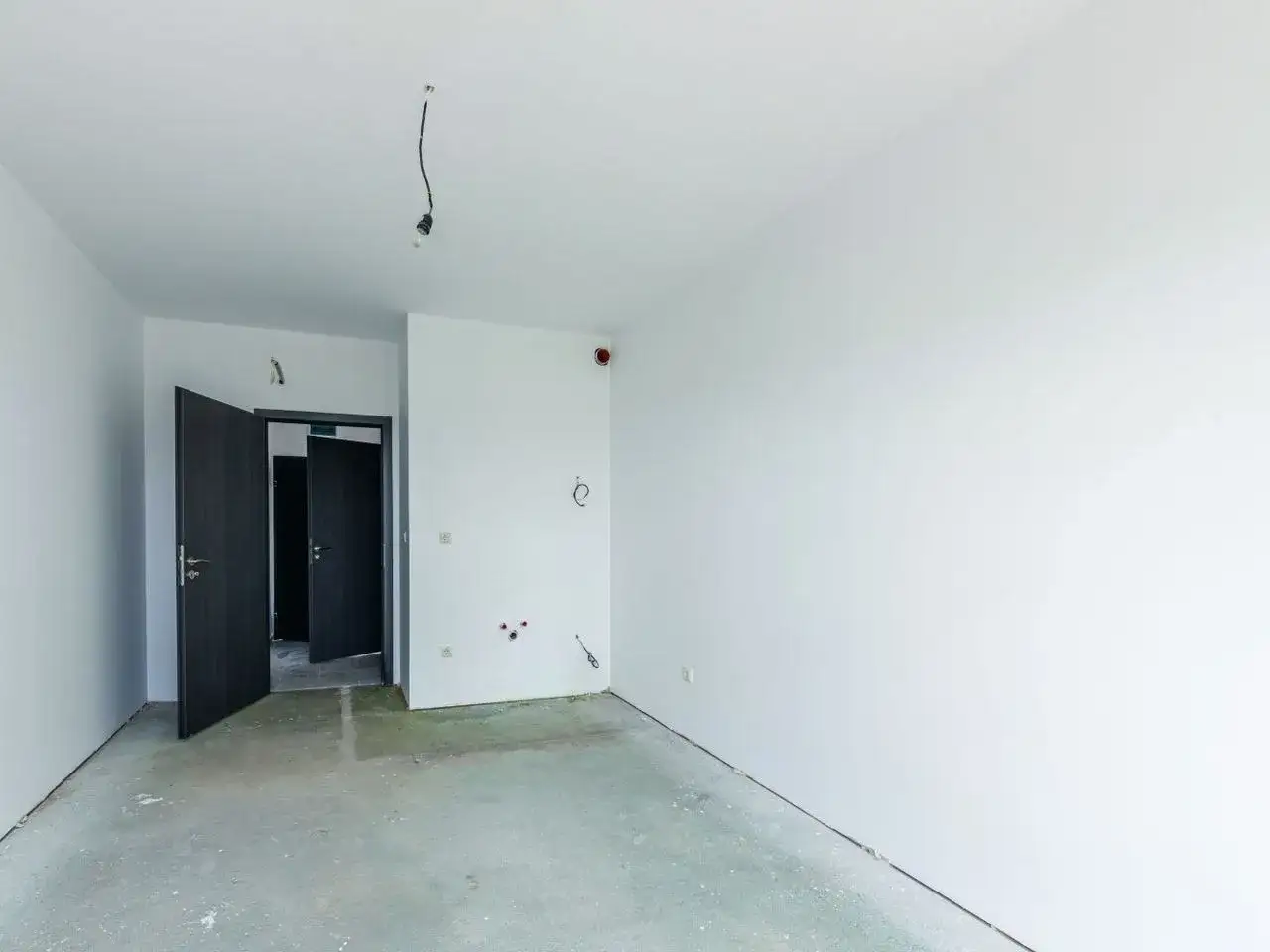 Puste pomieszczenie w stanie deweloperskim, z betonową podłogą, białymi ścianami i otwartymi drzwiami.