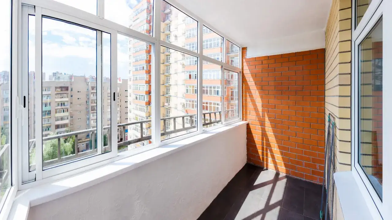 Zabudowana loggia z widokiem na blokowisko. Jasne okna i ceglana ściana tworzą przytulne miejsce, niczym dodatkowy balkon.