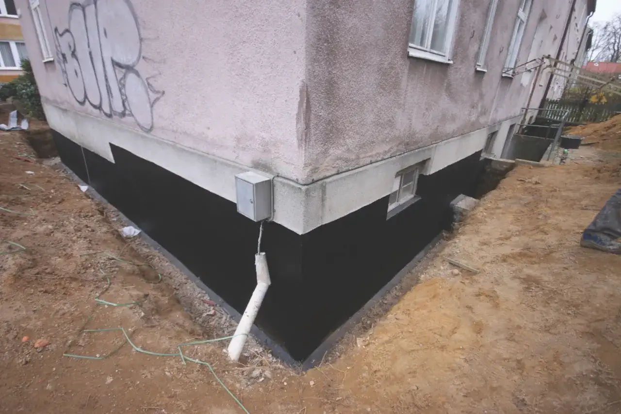 Prace przy izolacji fundamentów w starym domu. Czarna izolacja chroni mury przed wilgocią.