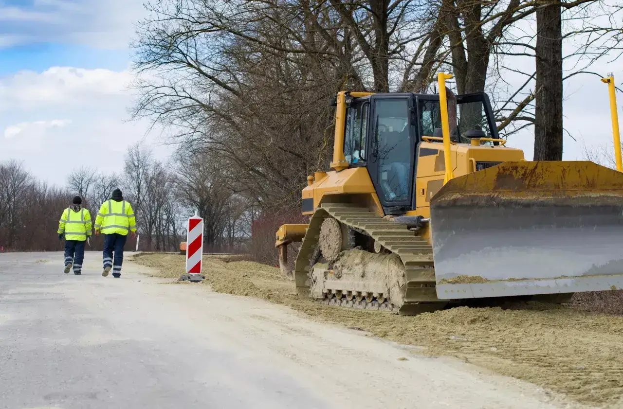 Dwóch robotników w kamizelkach odblaskowych idzie obok spychacza. Trwa praca związana z opłatą adiacencką.