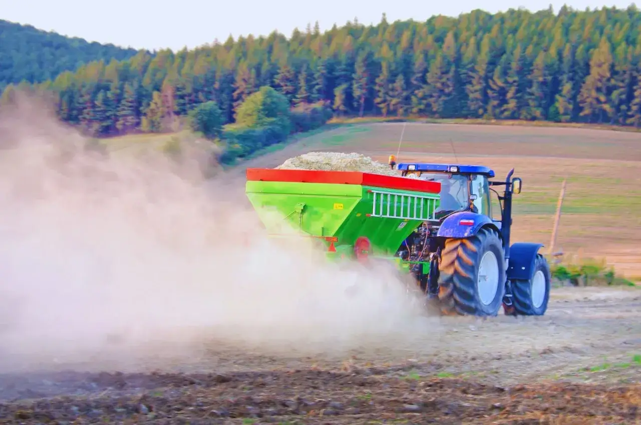 Traktor rozsiewa wapno palone na polu, tworząc chmurę pyłu. W tle las i wzgórza.