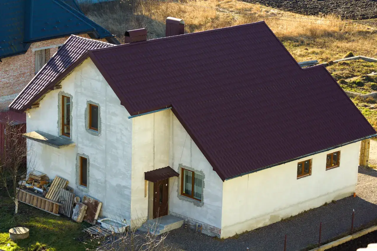 Dom w budowie na działce siedliskowej, z ciemnobordową, falistą dachówką i jasnymi ścianami. Wokół materiały budowlane.