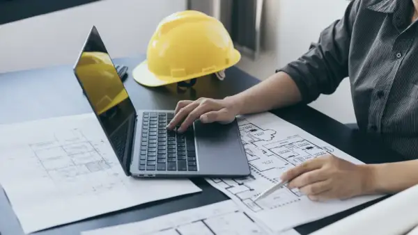 Architekt analizuje plany, obliczając powierzchnię użytkową budynku na laptopie. Obok leży żółty kask.