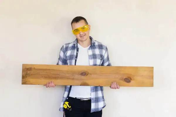 Mężczyzna w okularach ochronnych trzyma drewnianą deskę, która może posłużyć jako panele podłogowe na ścianie.