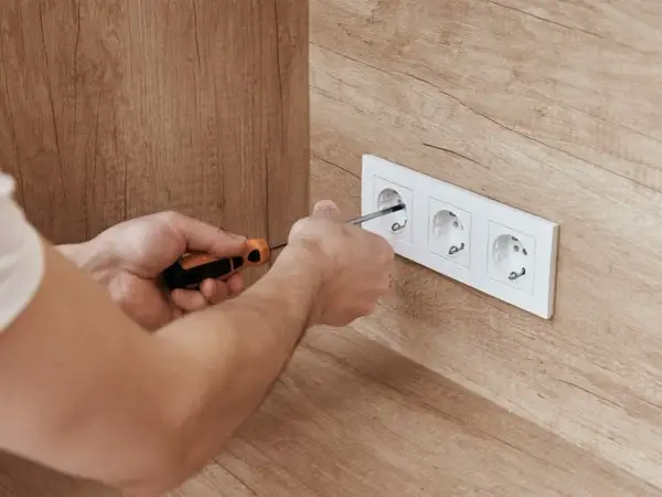 Ręce majsterkowicza dokręcają śrubki w białej ramce z trzema gniazdkami, zapewniając bezpieczne napięcie w gniazdku.