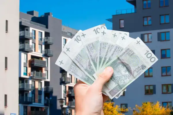 Dłoń trzyma polskie banknoty 100 zł na tle nowoczesnych bloków mieszkalnych. Szybka sprzedaż mieszkania przed upływem 5 lat.