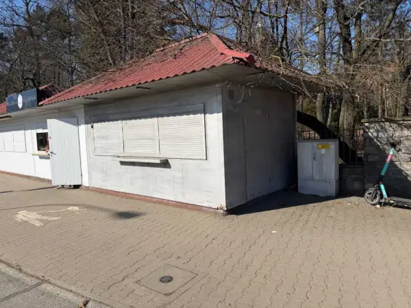 Mały lokal użytkowy z czerwoną, falistą dachem i białymi okiennicami. Obok stoi hulajnoga elektryczna.