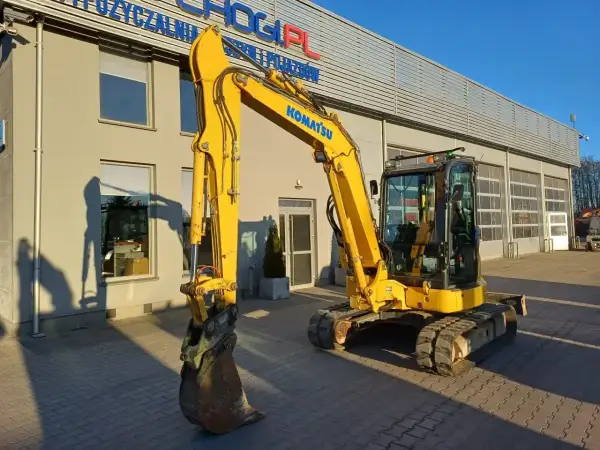 Żółta koparka Komatsu przed budynkiem. Zastanawiasz się, ile kosztuje godzina pracy koparki? Sprawdź ofertę wypożyczalni!